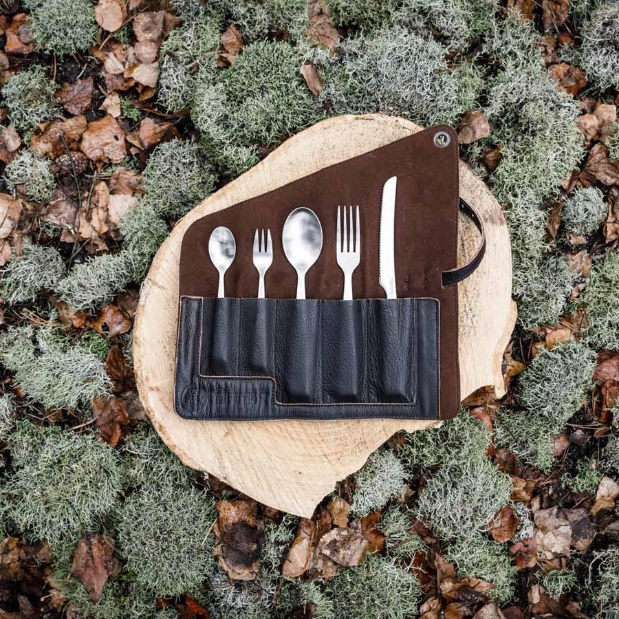 Set of cutlery in a leather case on a wooden surface surrounded by natural elements.